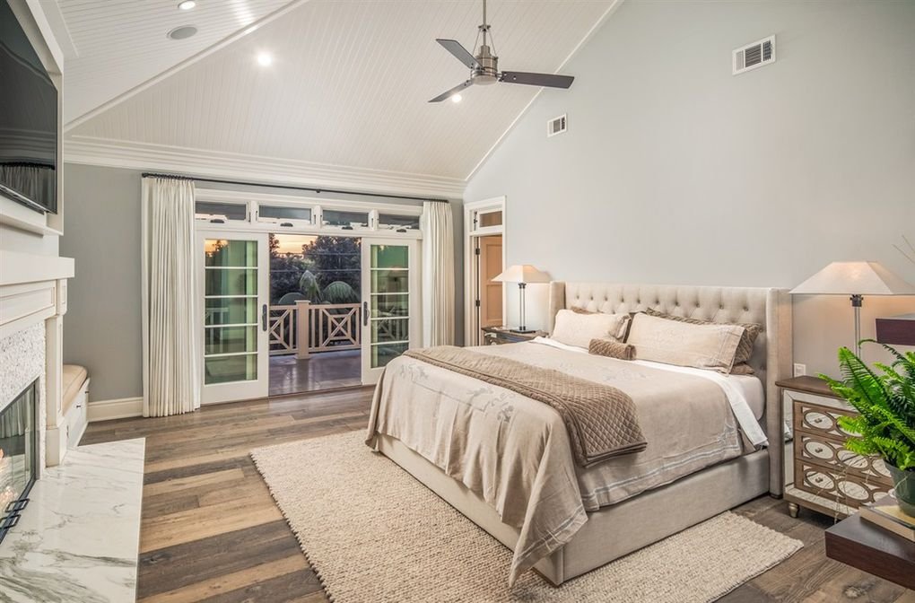 A spacious bedroom with a large bed, nightstands with lamps, sliding glass doors leading to a balcony, and a vaulted ceiling with a ceiling fan.