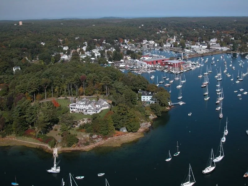 Aerial view of a coastal town with numerous sailboats docked in a marina, surrounded by lush greenery and residential houses.