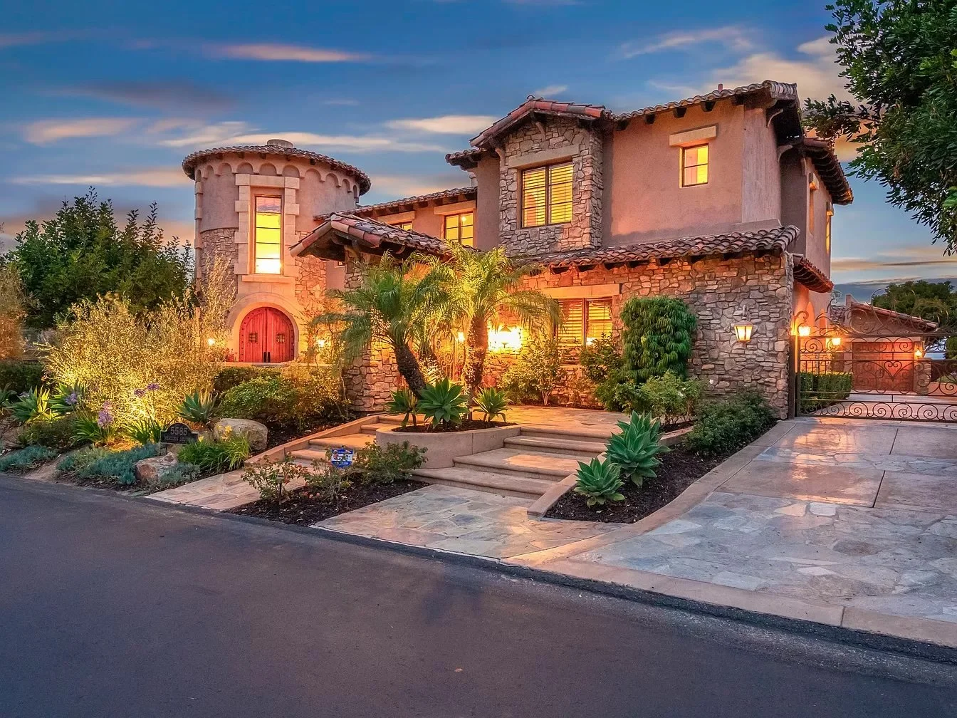 A large, stone and stucco house with warm lighting, a curved chimney, and a tiled roof, set against a twilight sky with a front yard featuring desert plants and a stone pathway.