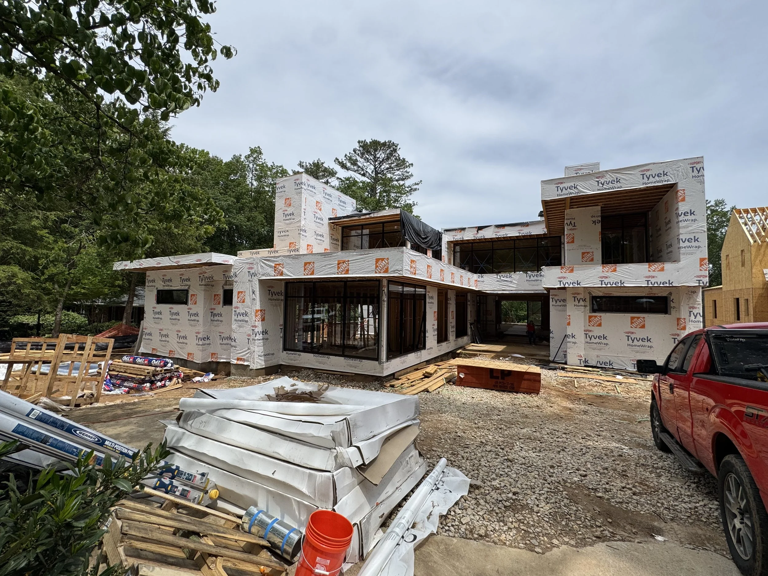 Construction site with a partially built house wrapped in Tyvek HomeWrap, surrounded by construction materials, debris, and parked cars, with trees and a cloudy sky in the background.