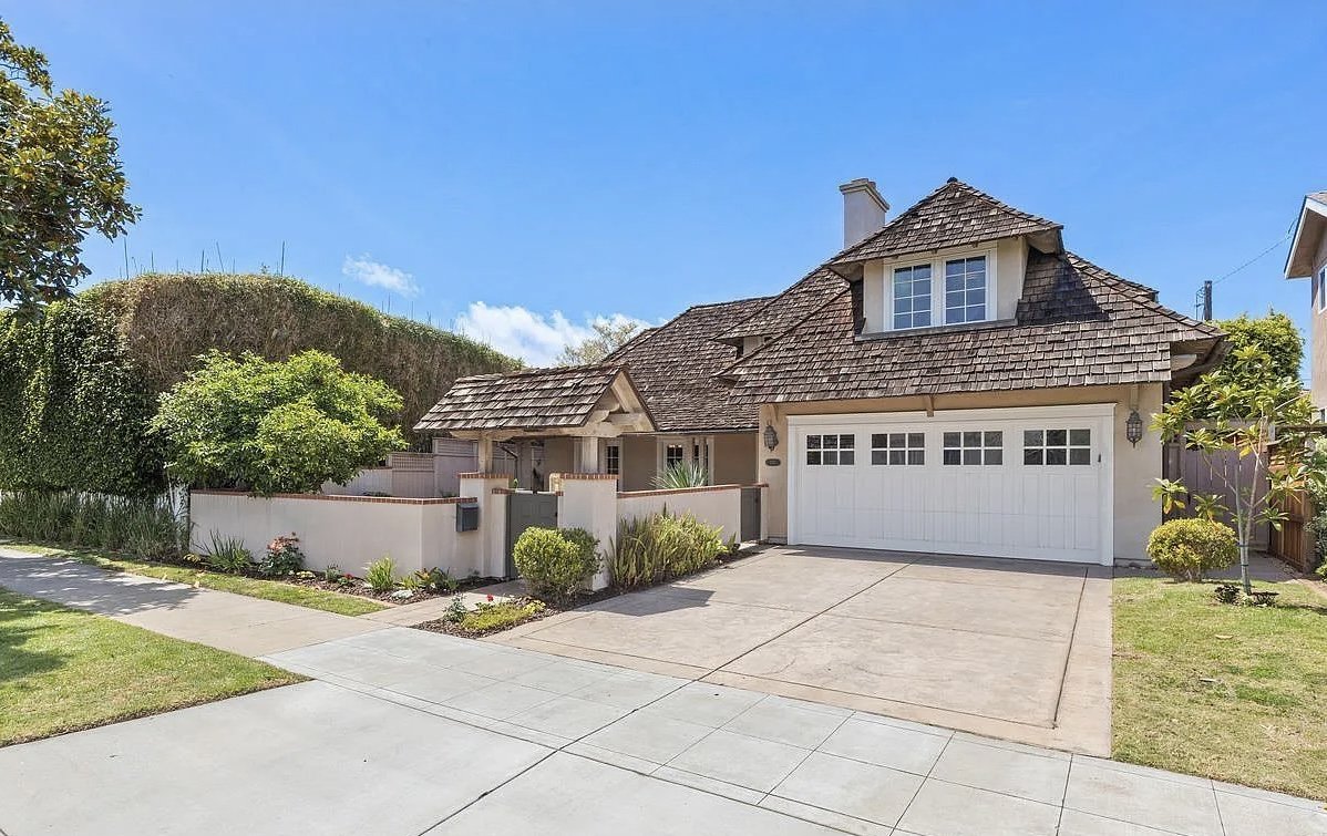 A house with a white garage door, tiled roof, and a small front yard with bushes and plants, under a blue sky.