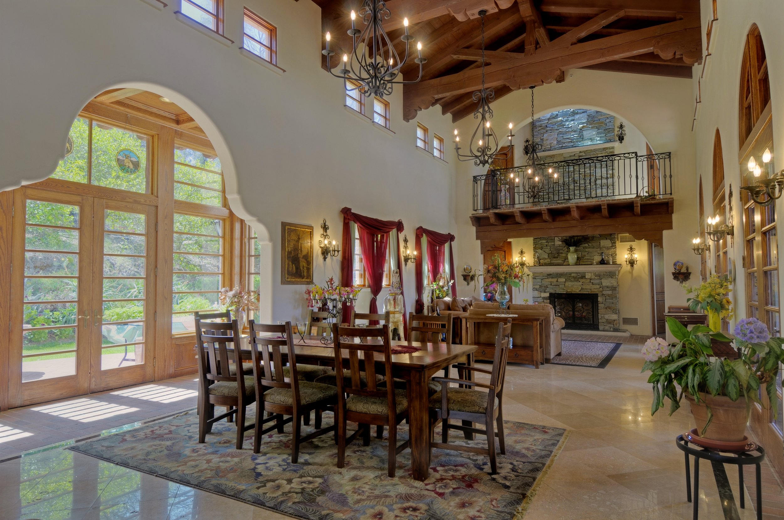 A spacious living room with large windows, wooden ceiling beams, and a stone fireplace. There are chandeliers hanging from the ceiling, and red curtains adorn the windows. The room is decorated with flowers and artworks, with a dining table and chair