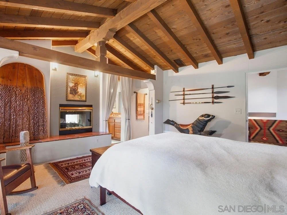 A cozy bedroom with a wooden ceiling, white walls, and rustic decor. It features a white bed, a small fireplace, a wooden chair, and mounted antique oars and a carved wooden fish on the wall.