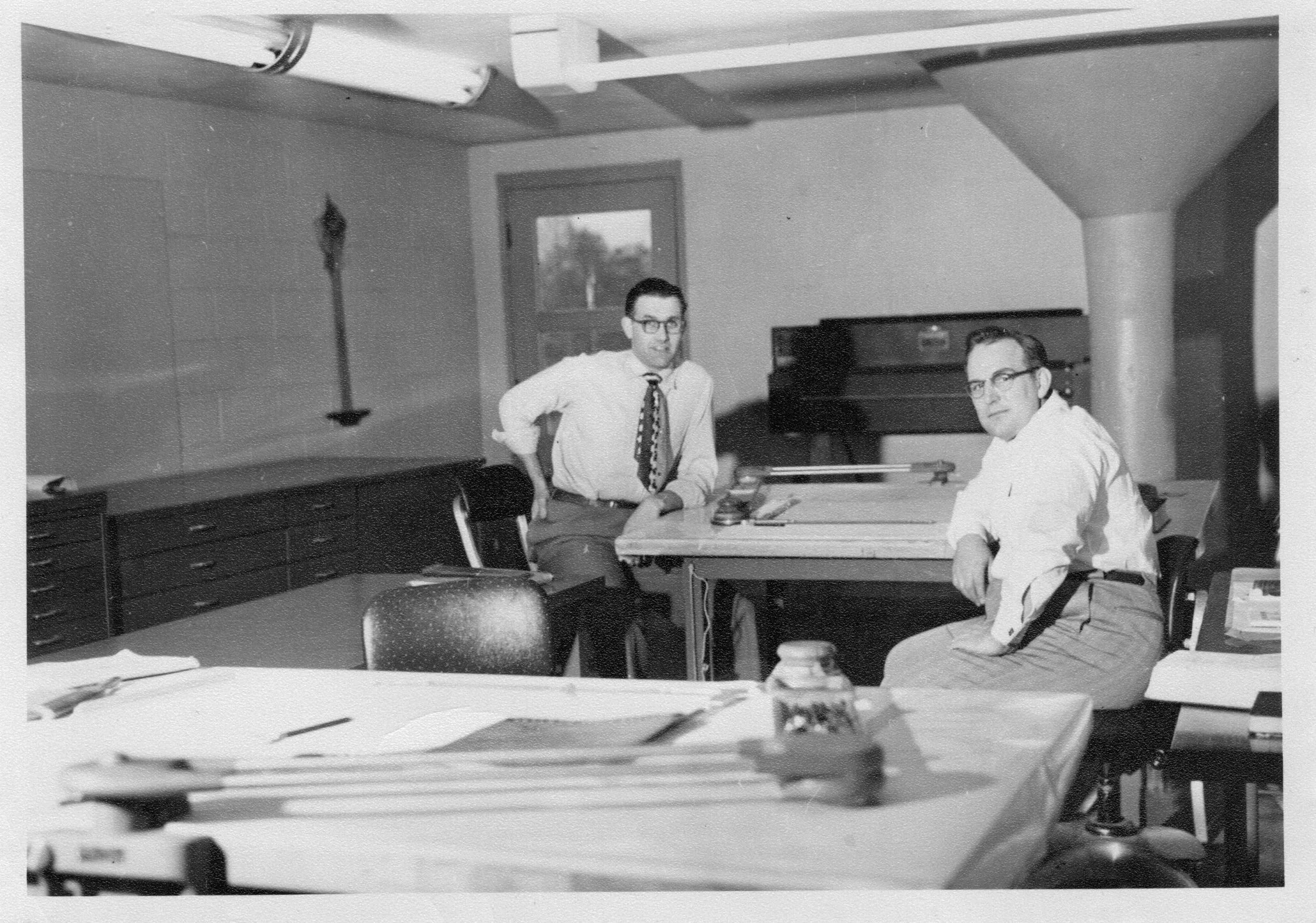Two men with glasses sitting at a desk in an office, one standing and the other seated, with shelves and a window in the background.