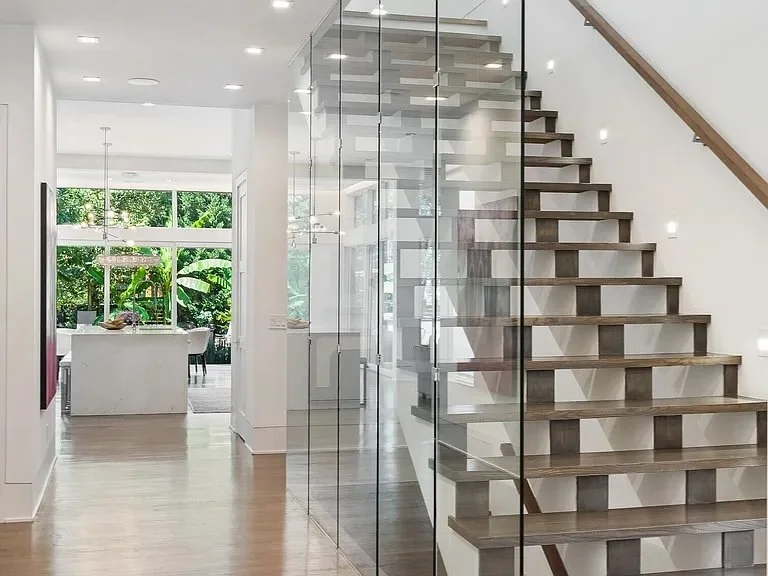 Modern interior with staircase, glass railing, and a view into a bright dining area with large windows and lush greenery outside.