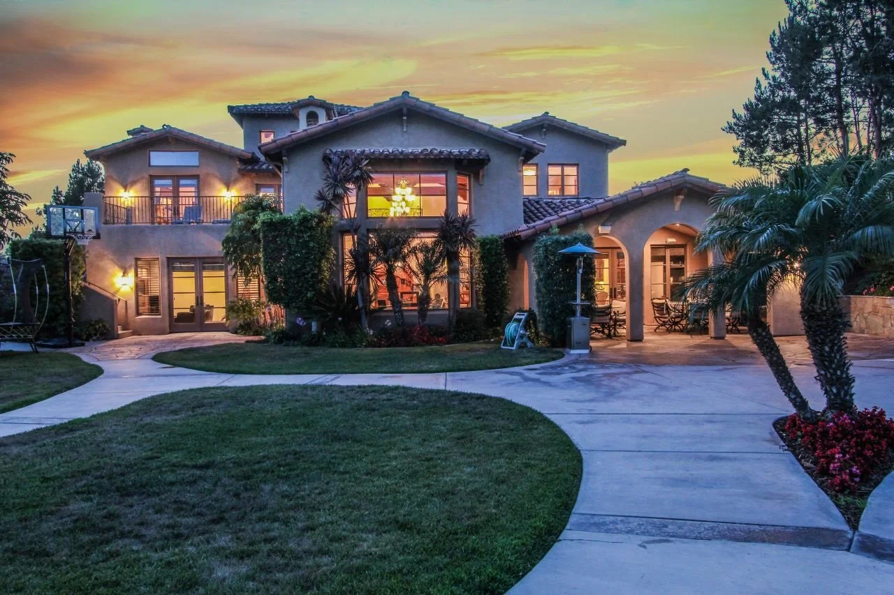 A large, two-story house with a stucco exterior, multiple large windows, and a tiled roof during sunset. The backyard features a curved concrete patio, a basketball hoop, palm trees, and outdoor furniture.