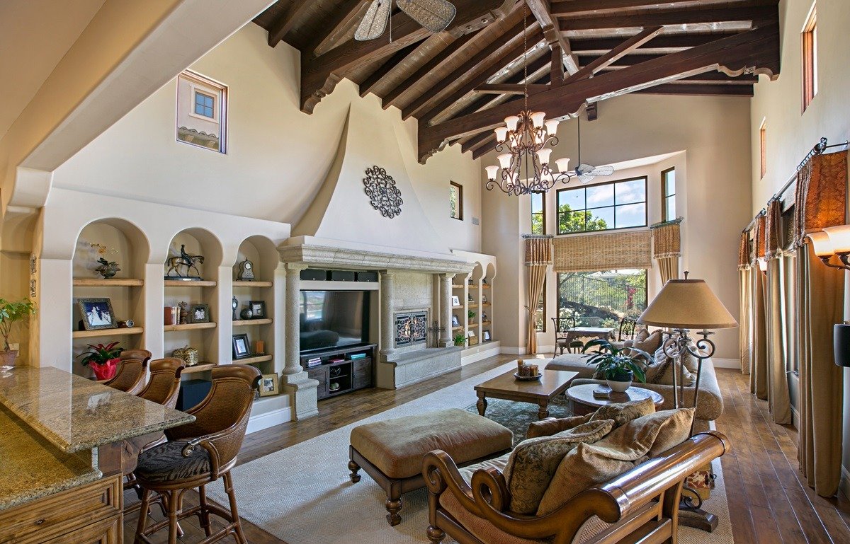 Living room with high vaulted ceiling, wooden beams, chandelier, large bay window, beige sofa with matching armchair, ottoman, coffee table, built-in shelves with decorative items, television, and large curtains.