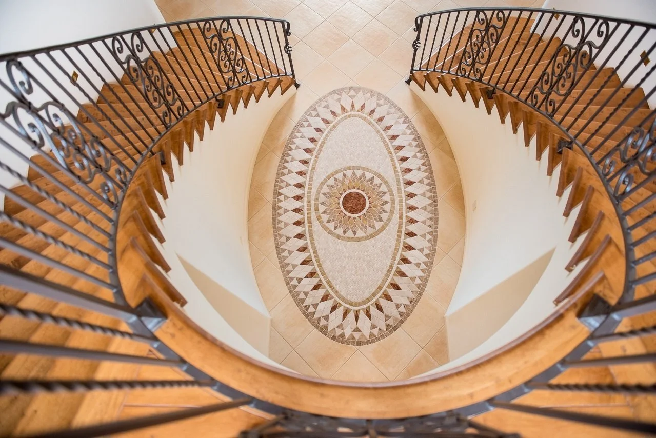 View looking down at a spiral staircase with wooden steps and black wrought iron railing, overlooking a patterned tile floor below.