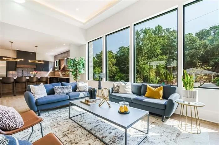 Modern living room with large windows showing trees outside, two blue sofas with pillows, a white coffee table, and a kitchen in the background.