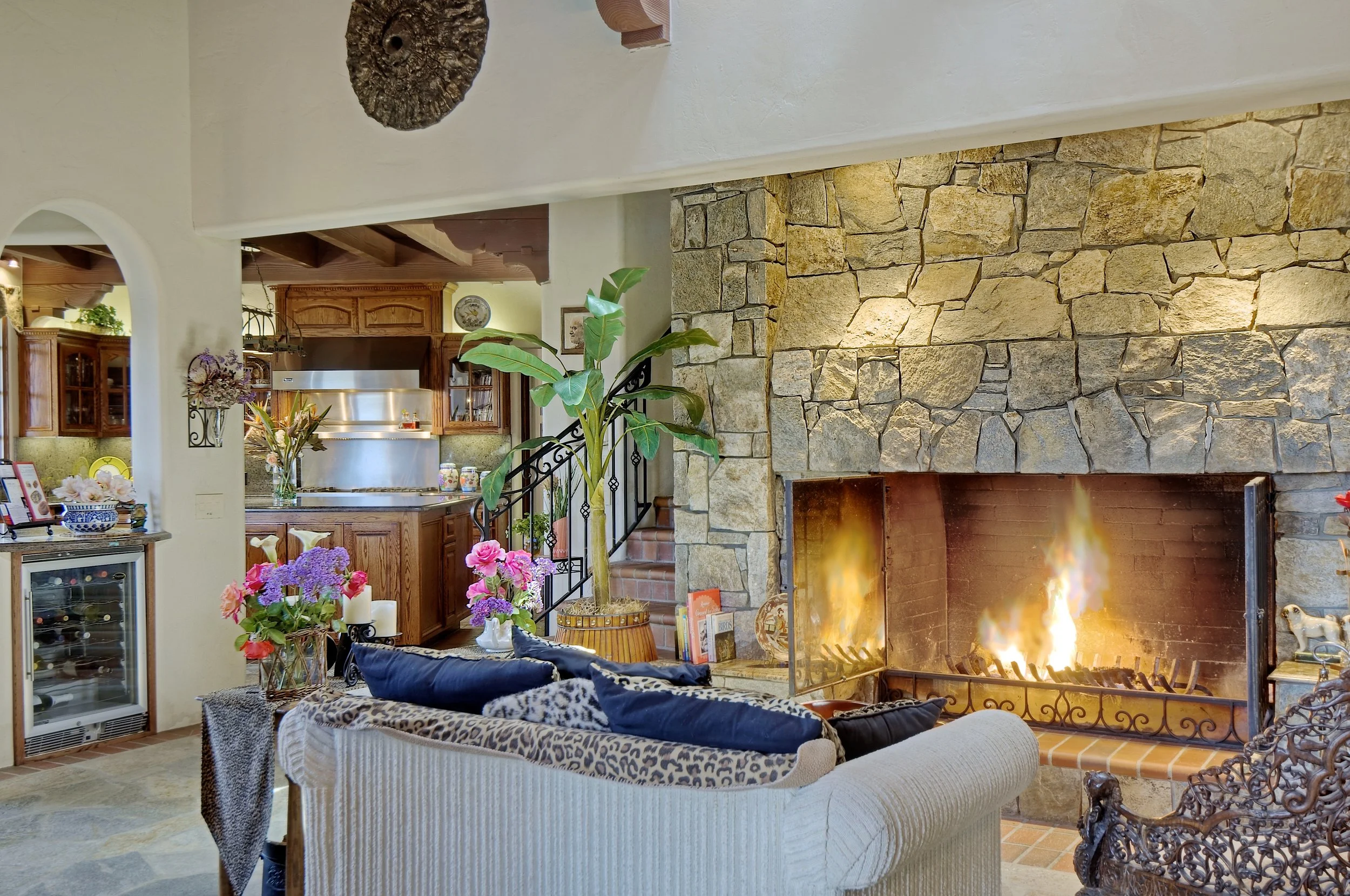 Living room with a lit fireplace, stone mantel, patterned and plain couches, plants, and decorative flowers, in a cozy, rustic style.
