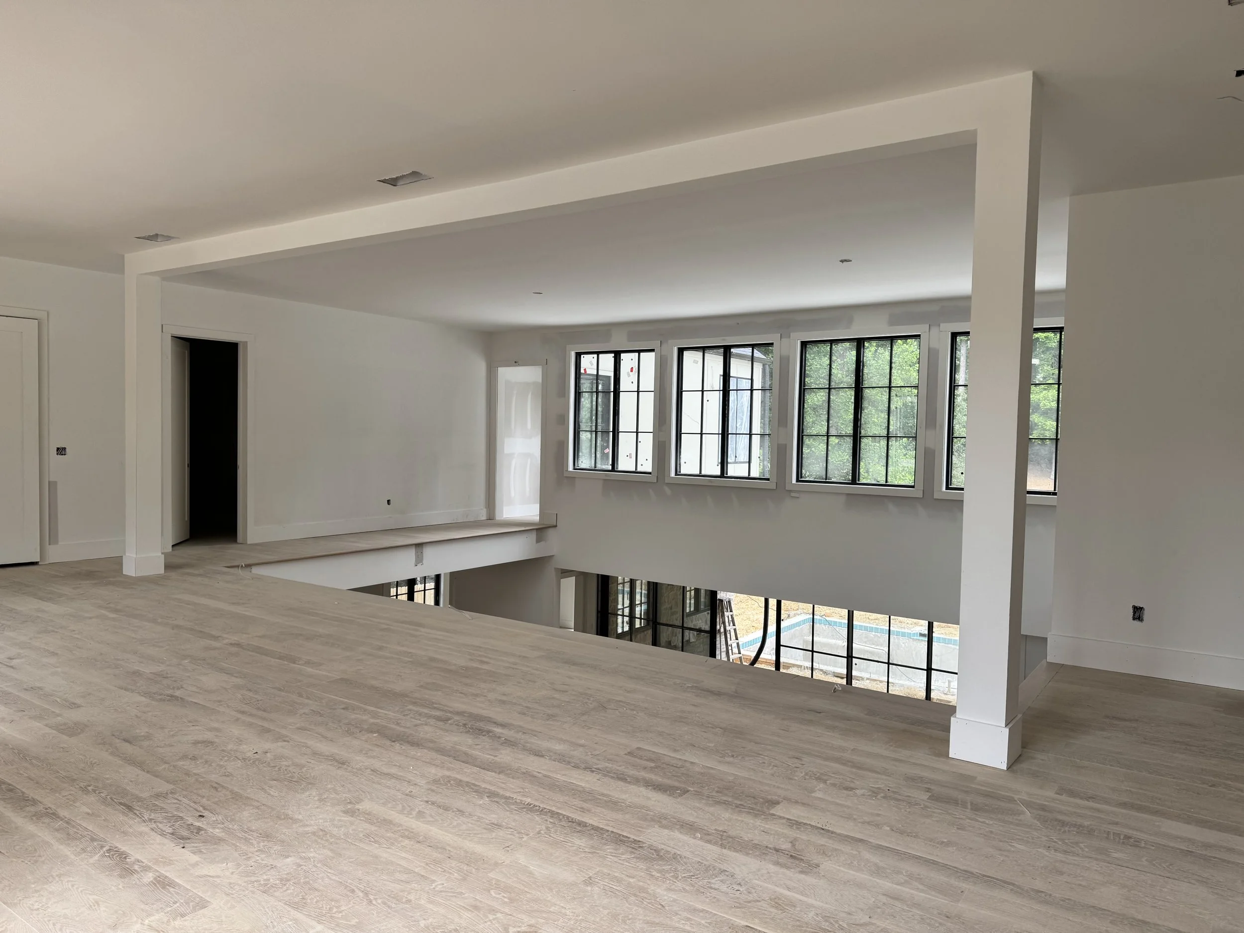 An unfinished modern open-concept living room with large windows, light wood flooring, and white walls and ceiling. There is a partial wall with a ledge and a staircase leading down, visible through an opening. Outside, there is a deck and greenery.