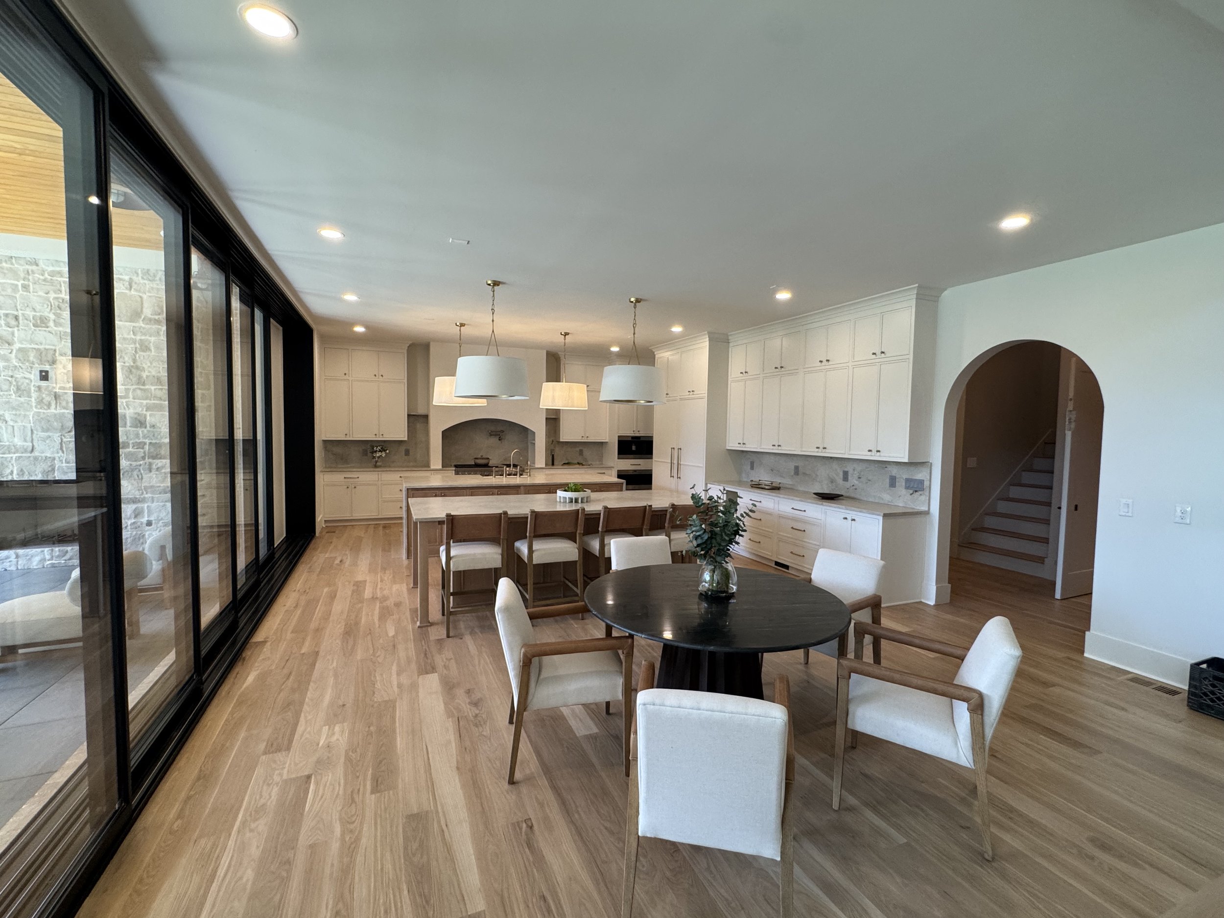 Open-concept kitchen and dining area with white cabinetry, pendant lighting, and a view of a sliding glass door leading outside. Wooden flooring and a round dining table with chairs are also visible.