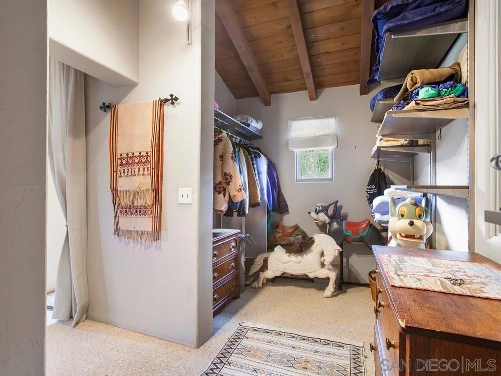 A small room with clothes on hanging racks, shelves with folded clothes and toys, a small window, and a rocking horse. There are wooden shelves and furniture, and a patterned rug on the carpeted floor.
