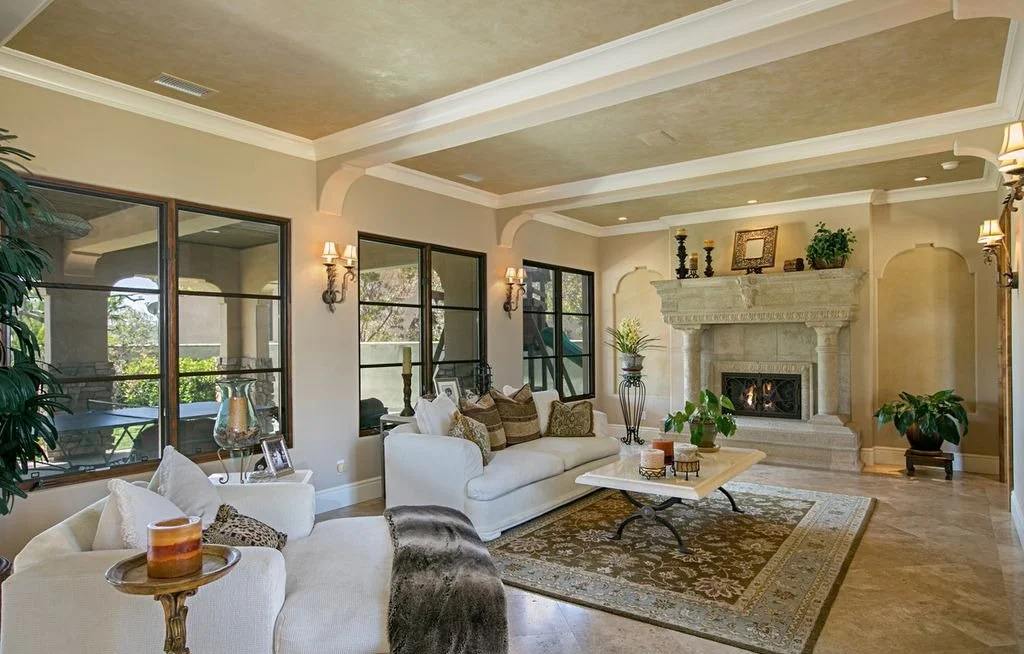 Elegant living room with white sofas, decorative pillows, a stone fireplace, large windows, potted plants, and a coffee table on a patterned rug.