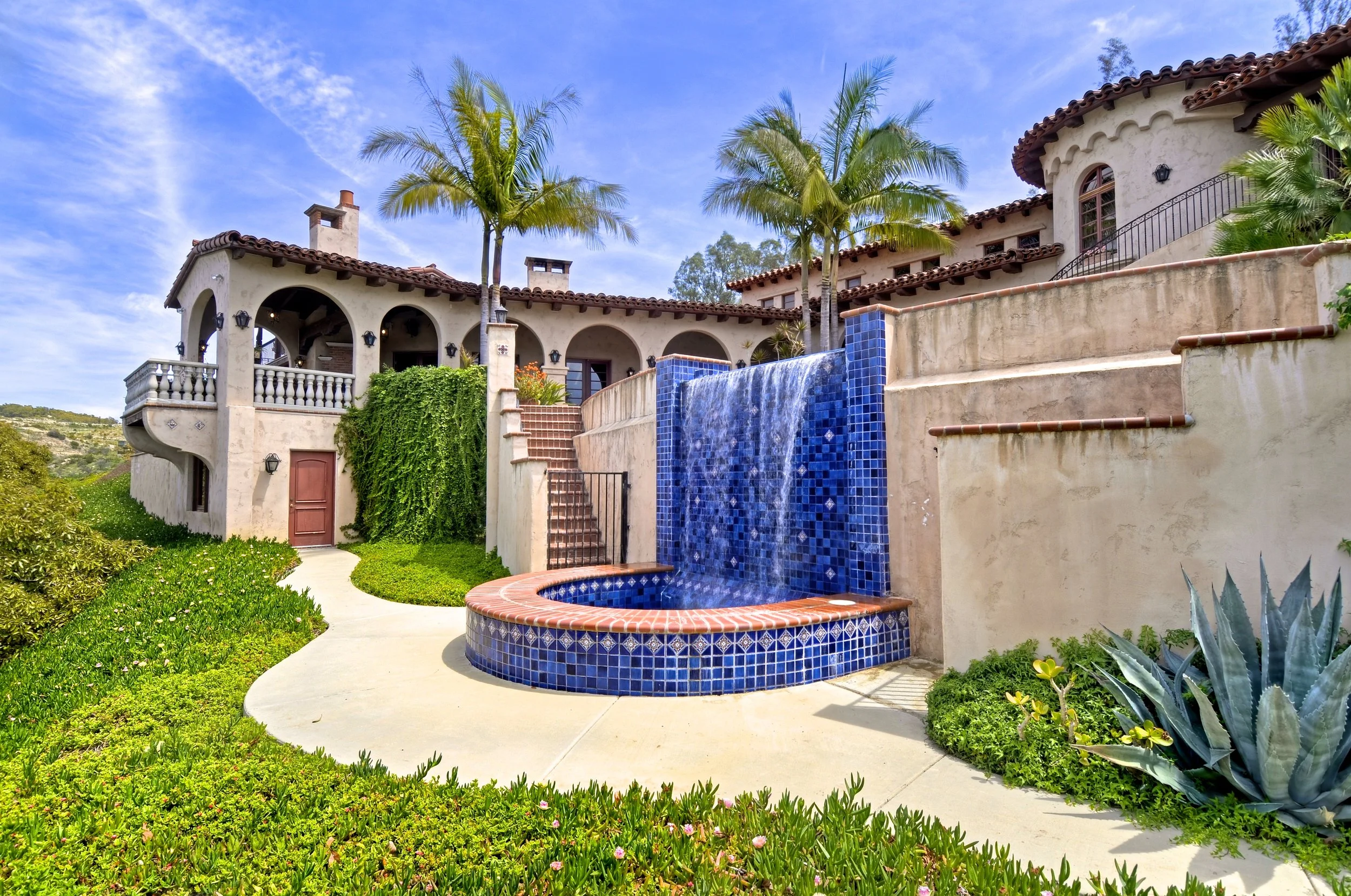 A Mediterranean-style house with a tiled roof, arched doorways, and a water fountain with blue tiles in the backyard, surrounded by lush green plants and palm trees.