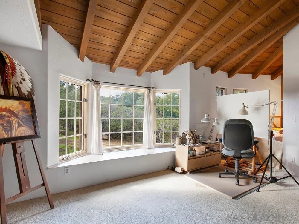 A bright room with large bay windows, wooden ceiling with exposed beams, a gray office chair, a wooden desk with lamps, a small shelf with art supplies, and a painting on an easel.