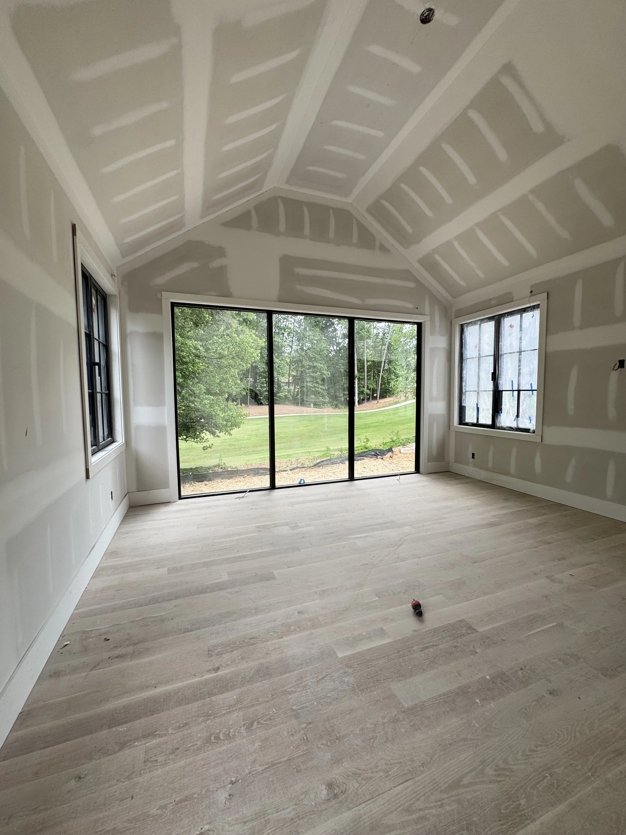 Room under construction with drywall taped and mudded, large windows looking outside at greenery, light-colored wooden flooring, some construction tools on the floor.