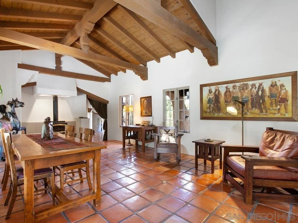 Living room with wooden ceiling beams, terracotta tile floor, white walls, wooden furniture including a dining table, chairs, side tables, and a leather armchair. Windows letting in natural light, framed artwork on the walls, and decorative items on 