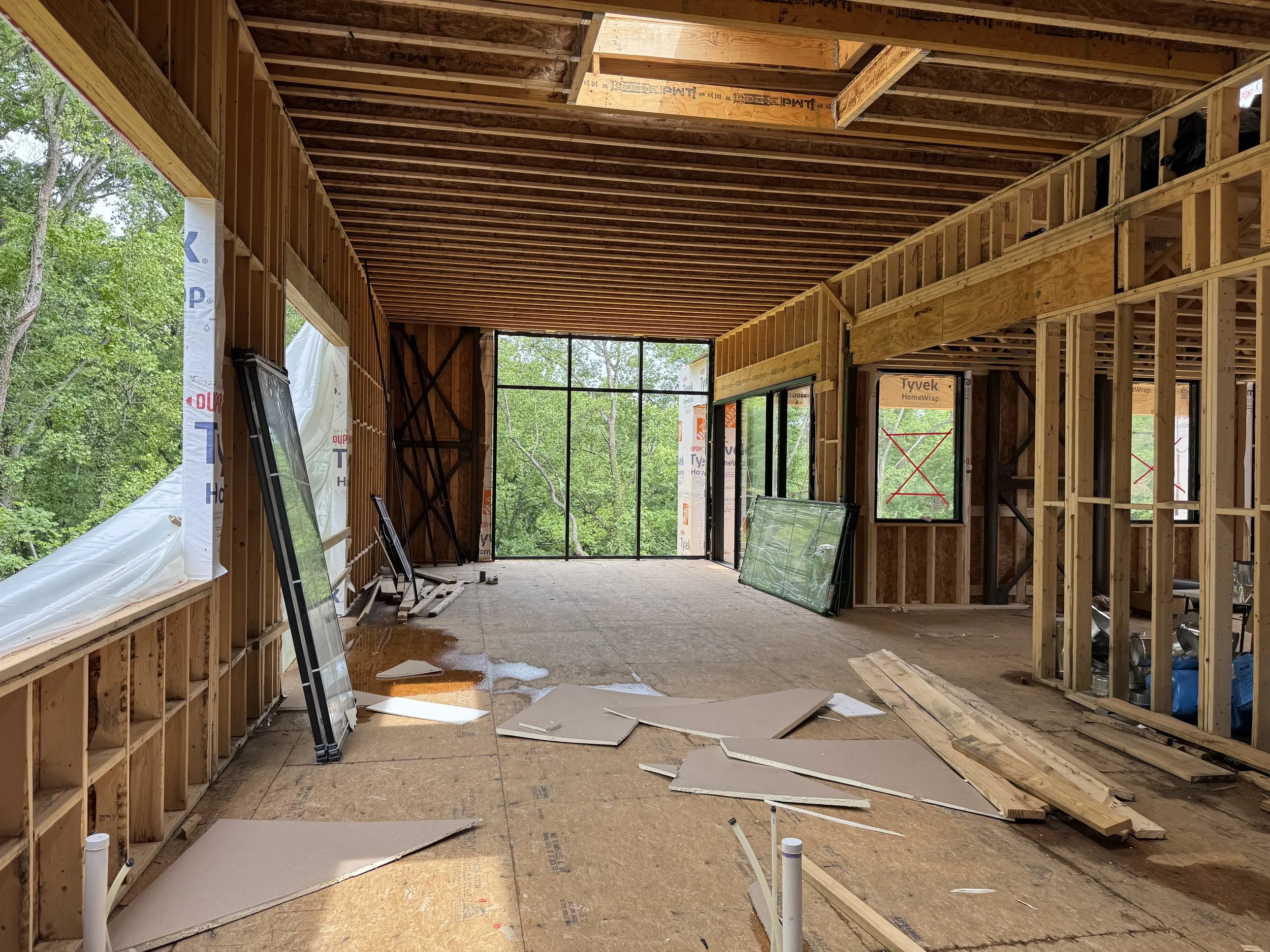 Interior view of a house under construction, showing exposed wooden framing, roof trusses, large windows, and construction debris on the floor.