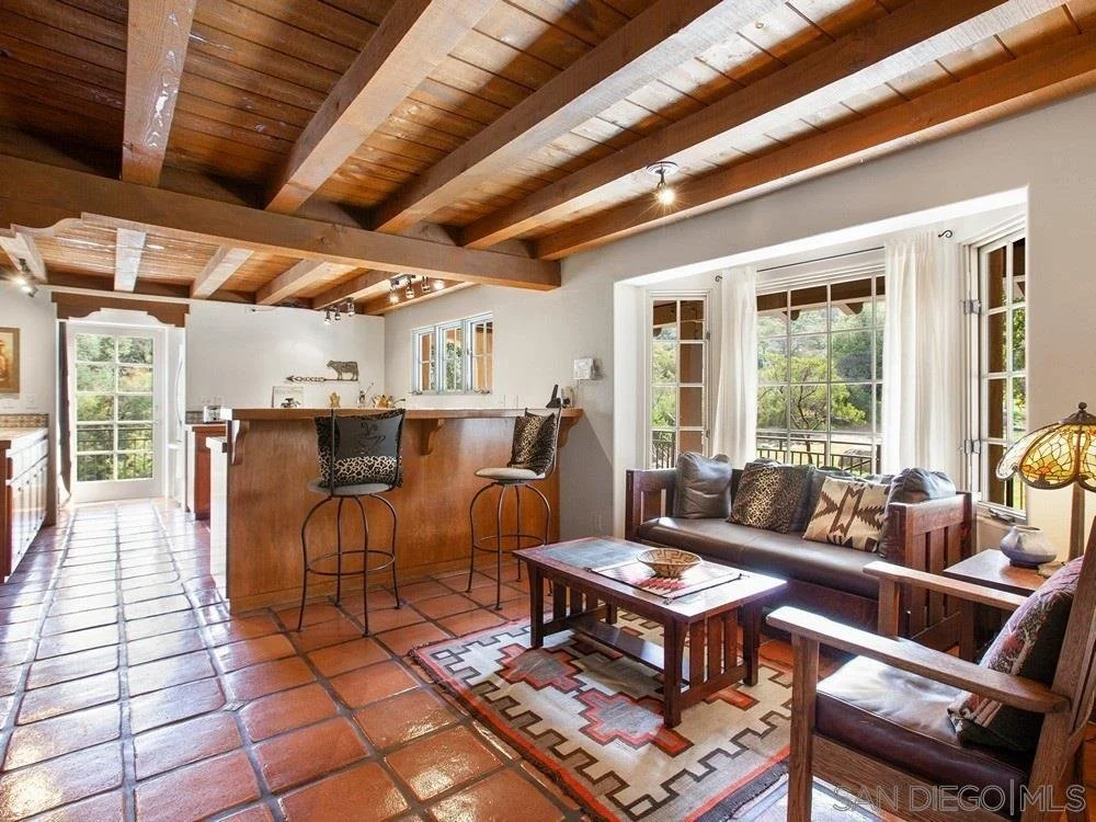 Living room with large windows, wooden ceiling beams, terracotta tile floor, and rustic furniture including a sofa, armchair, and coffee table.