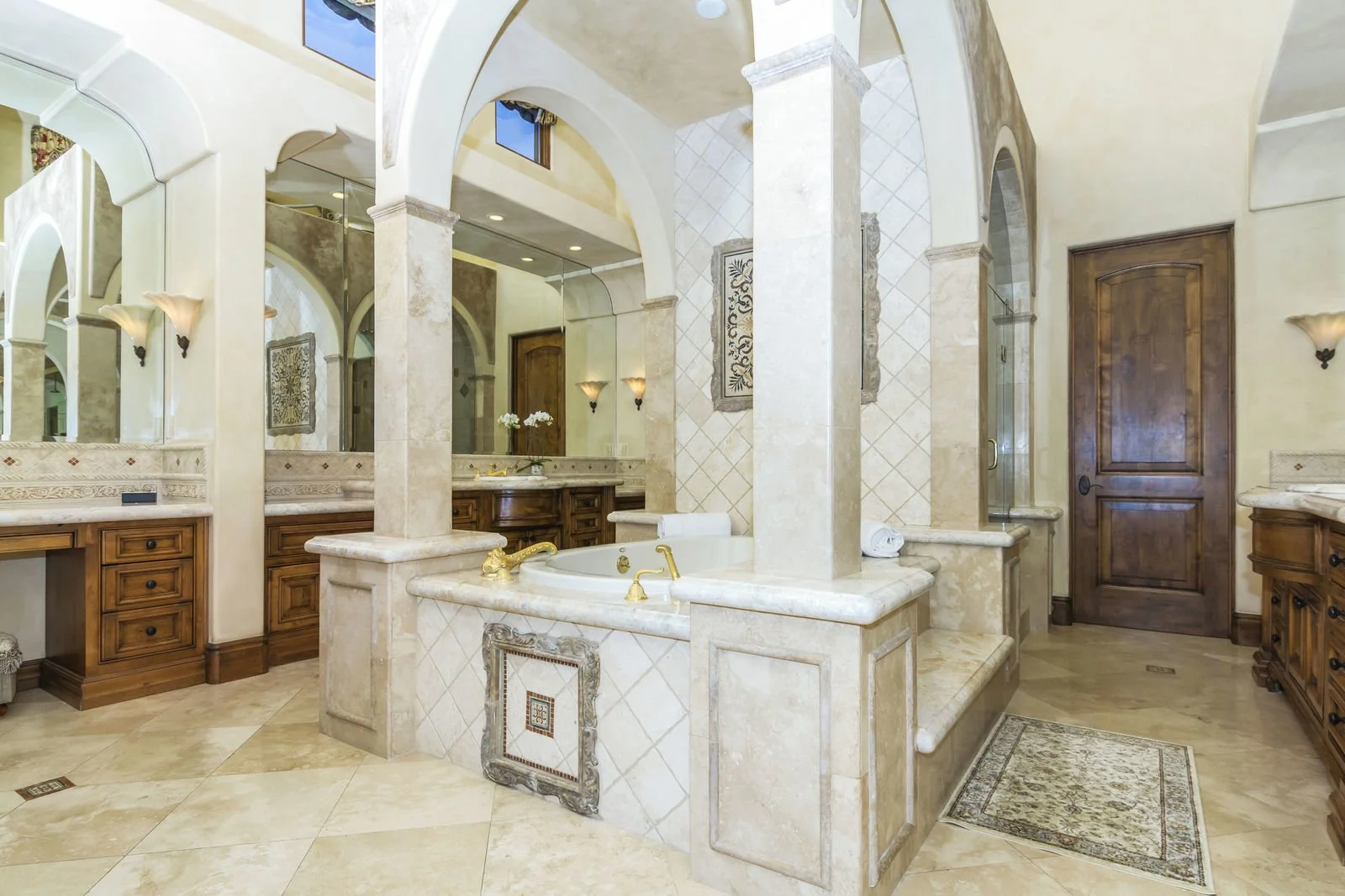 Luxurious bathroom with a large bathtub, marble countertops, wooden cabinets, mirrors, wall-mounted sconces, a glass-enclosed shower, decorative wall art, and a door.
