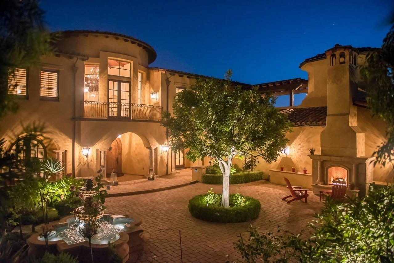 A well-lit courtyard of a Spanish-style house at night with a central tree, outdoor fire fireplace, seating area, and a water fountain.