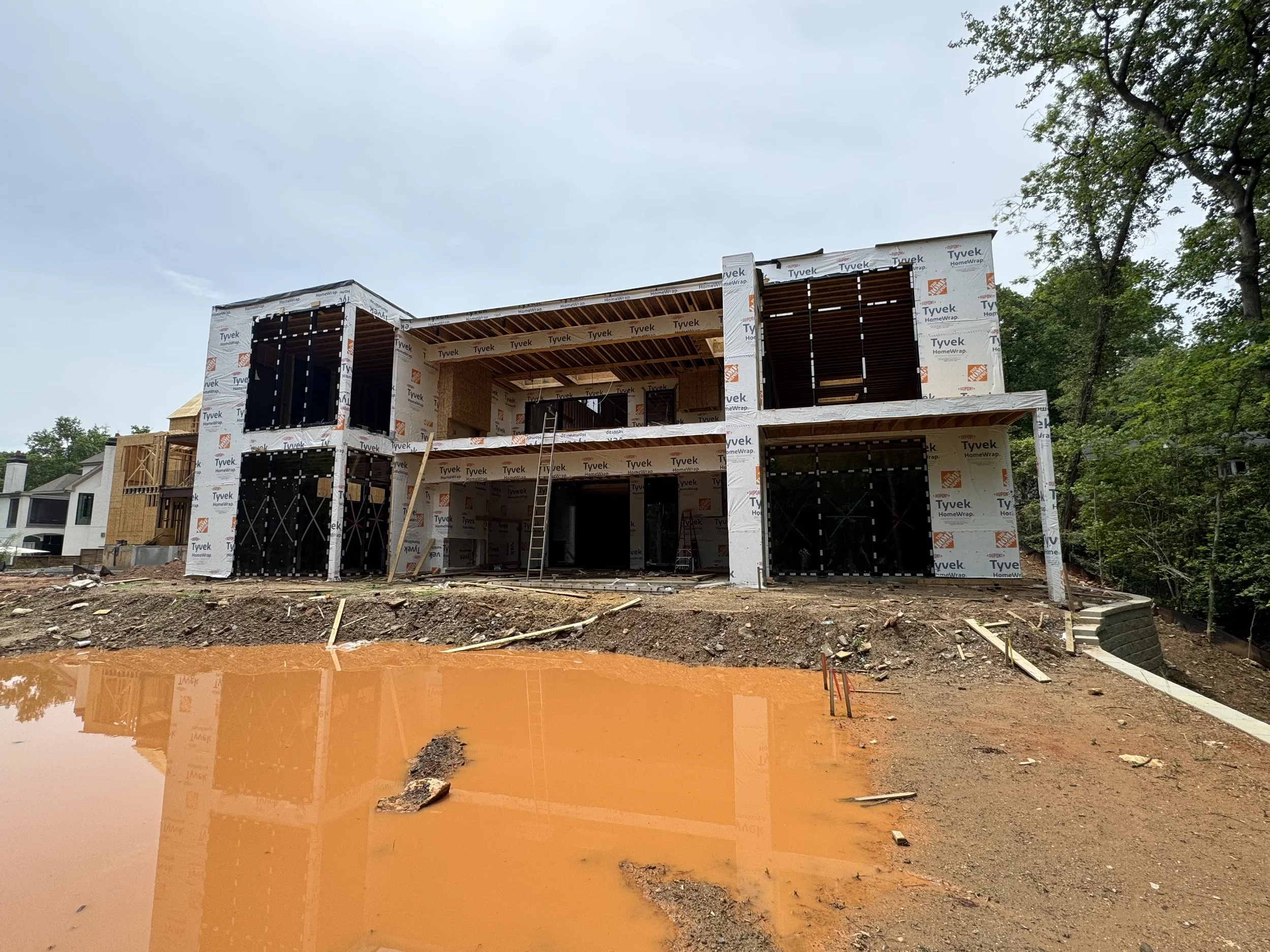 Construction site of a house with a partially built second floor, wrapped in Tyvek HomeWrap, surrounded by trees and a muddy ground with puddles of water.