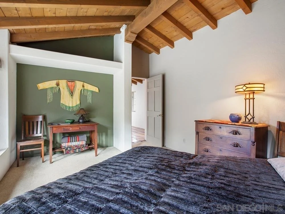 Bedroom with wooden ceiling, complete with a bed, dresser, table, and chair. Decorative textile hanging on wall and a lit table lamp on dresser.