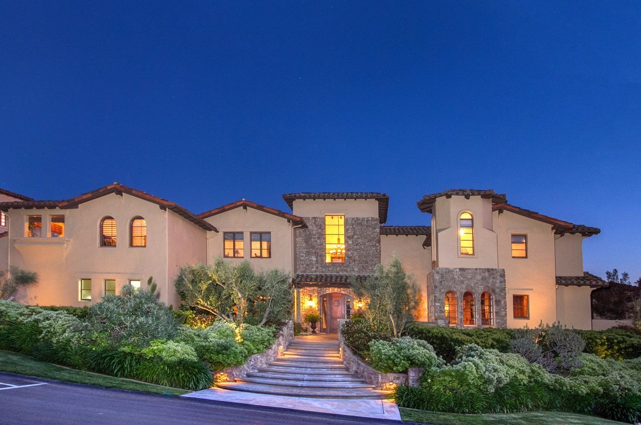 A large, elegant house with Mediterranean-style architecture, beige stucco walls, stone accents, and a terracotta tile roof illuminated at dusk, surrounded by well-maintained landscaping and greenery.