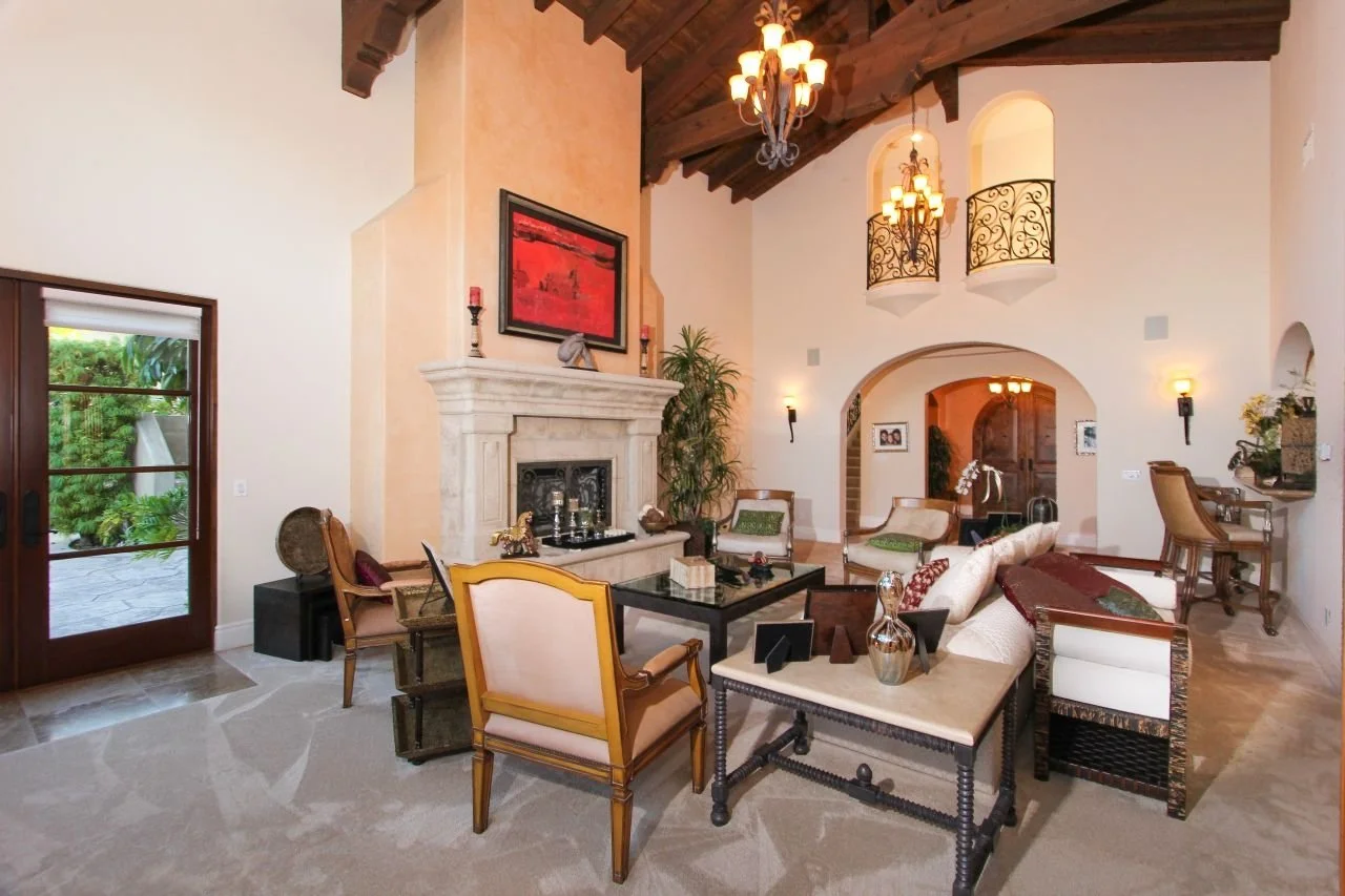 Spacious living room with high ceilings, wooden beams, and a large stone fireplace. Elegant furniture, chandeliers, and decorative accents create a warm, inviting atmosphere. Multiple armchairs and sofas are arranged around a coffee table near a slid