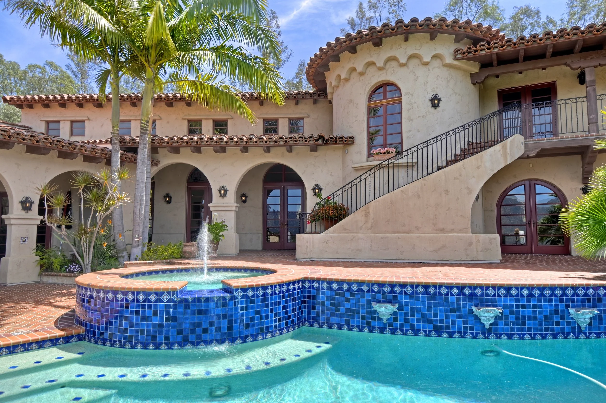 A luxurious two-story house with a Spanish-style architecture featuring stucco walls, arched doorways, and a red tile roof. A staircase leads up to the second floor balcony with an iron railing. In the foreground, there's a swimming pool with blue mo