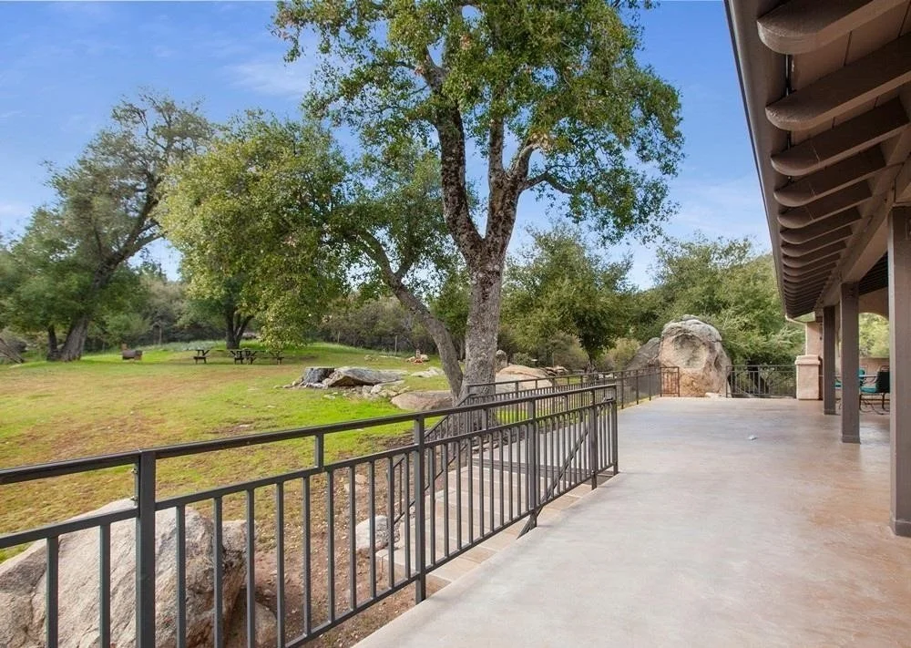 View of a spacious outdoor patio area with a metal railing, adjacent to a grassy yard with large trees and rocks under a clear blue sky.