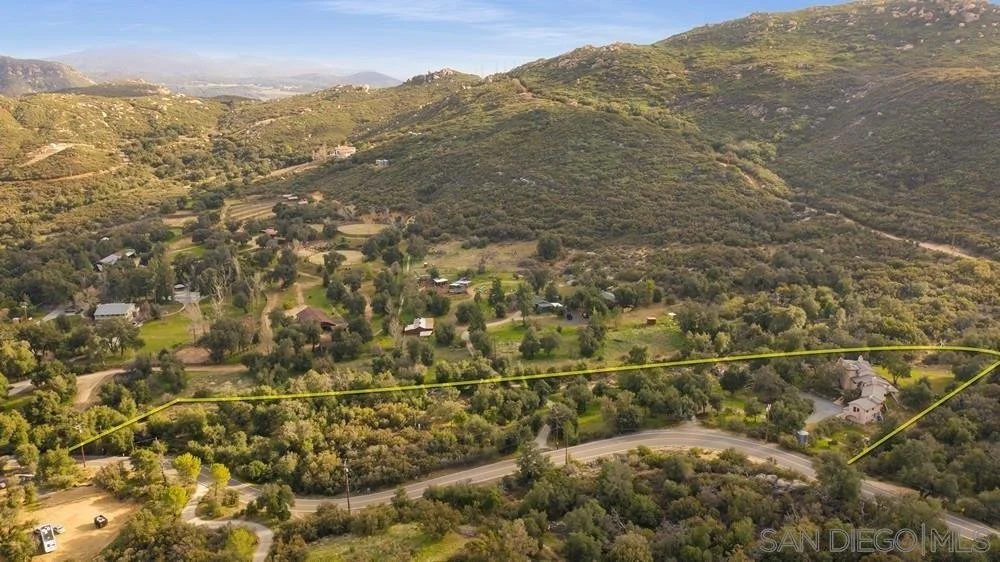 Aerial view of a hillside residential area with winding roads, houses, and lush green vegetation, surrounded by mountains under a clear sky.