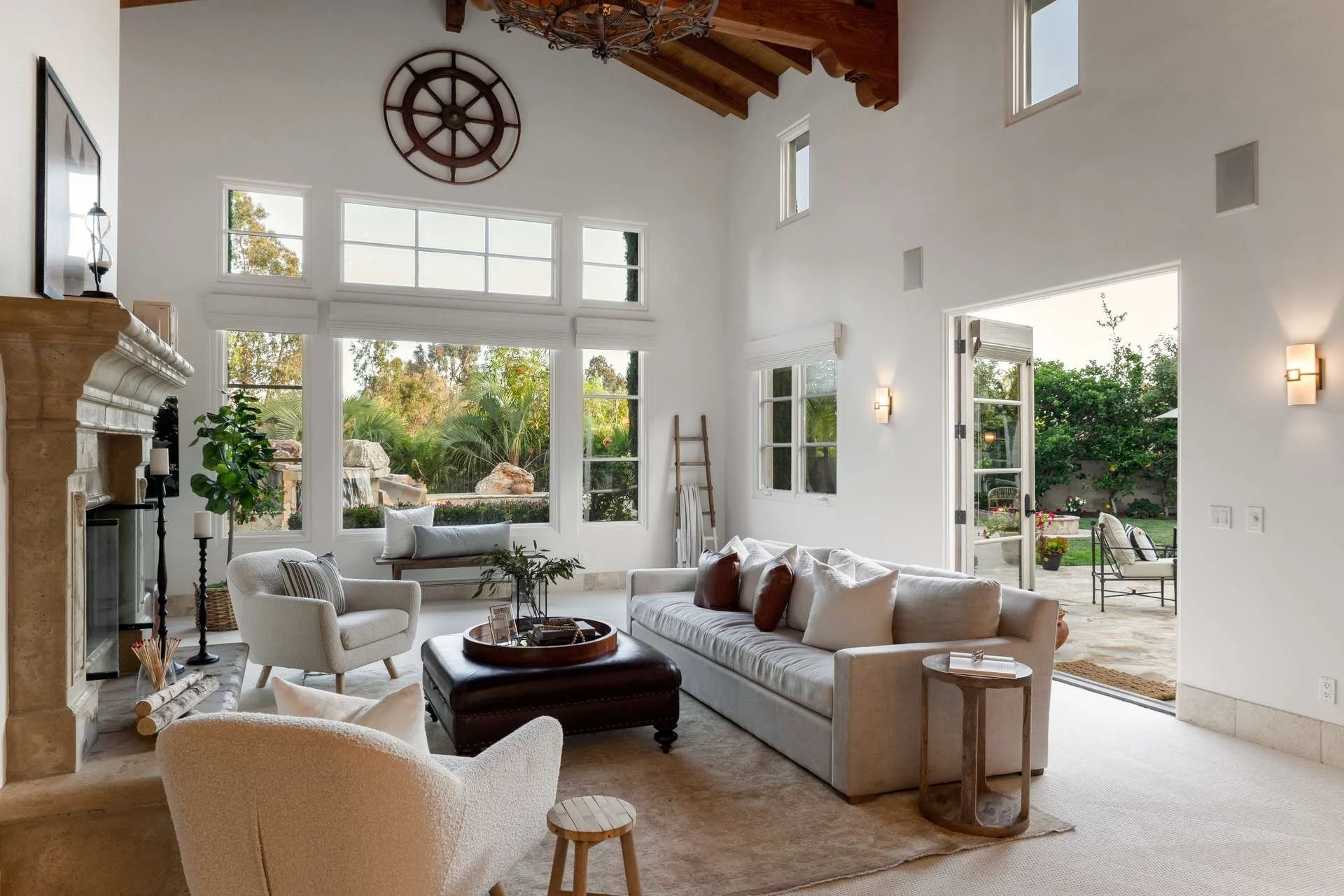 Bright living room with white walls and high ceiling, large windows and glass door opening to a garden patio, beige sofa and white armchairs, fireplace, and decor including a large wall clock, ladder, plants, and cushions.