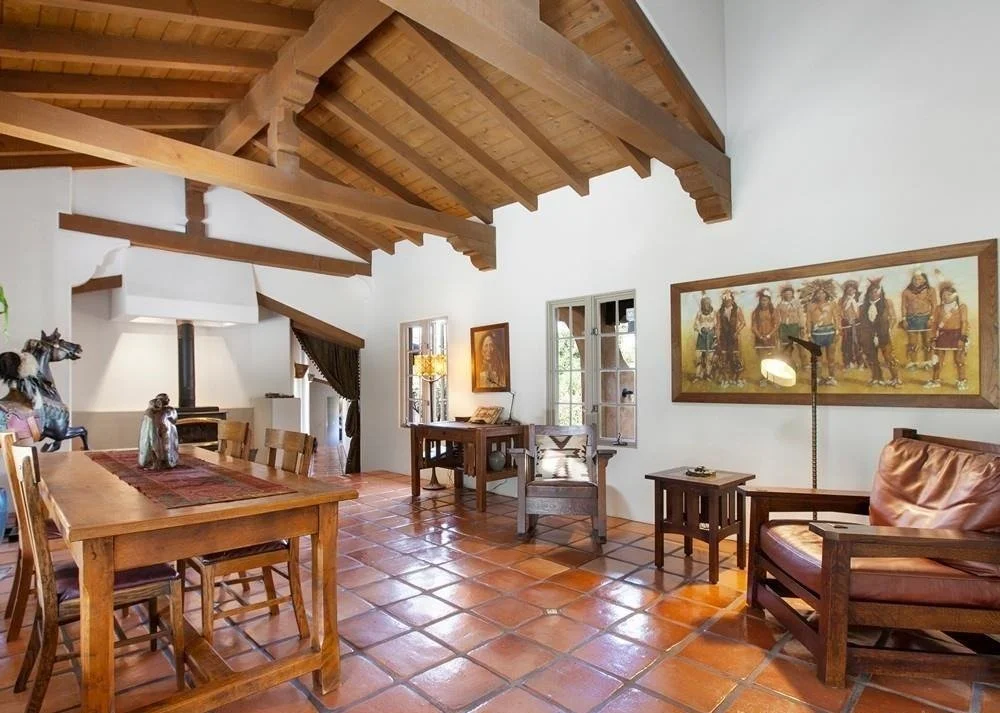 Living room with wooden ceiling beams, terracotta tile floor, and rustic furniture. Includes a leather armchair, several wooden dining chairs, decorative animal statues, a side table, and artwork on the walls.