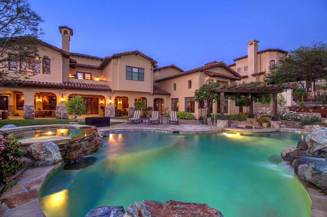 A residential-style backyard with a swimming pool, lounge chairs, trees, and a house with multiple windows and a patio area at dusk.
