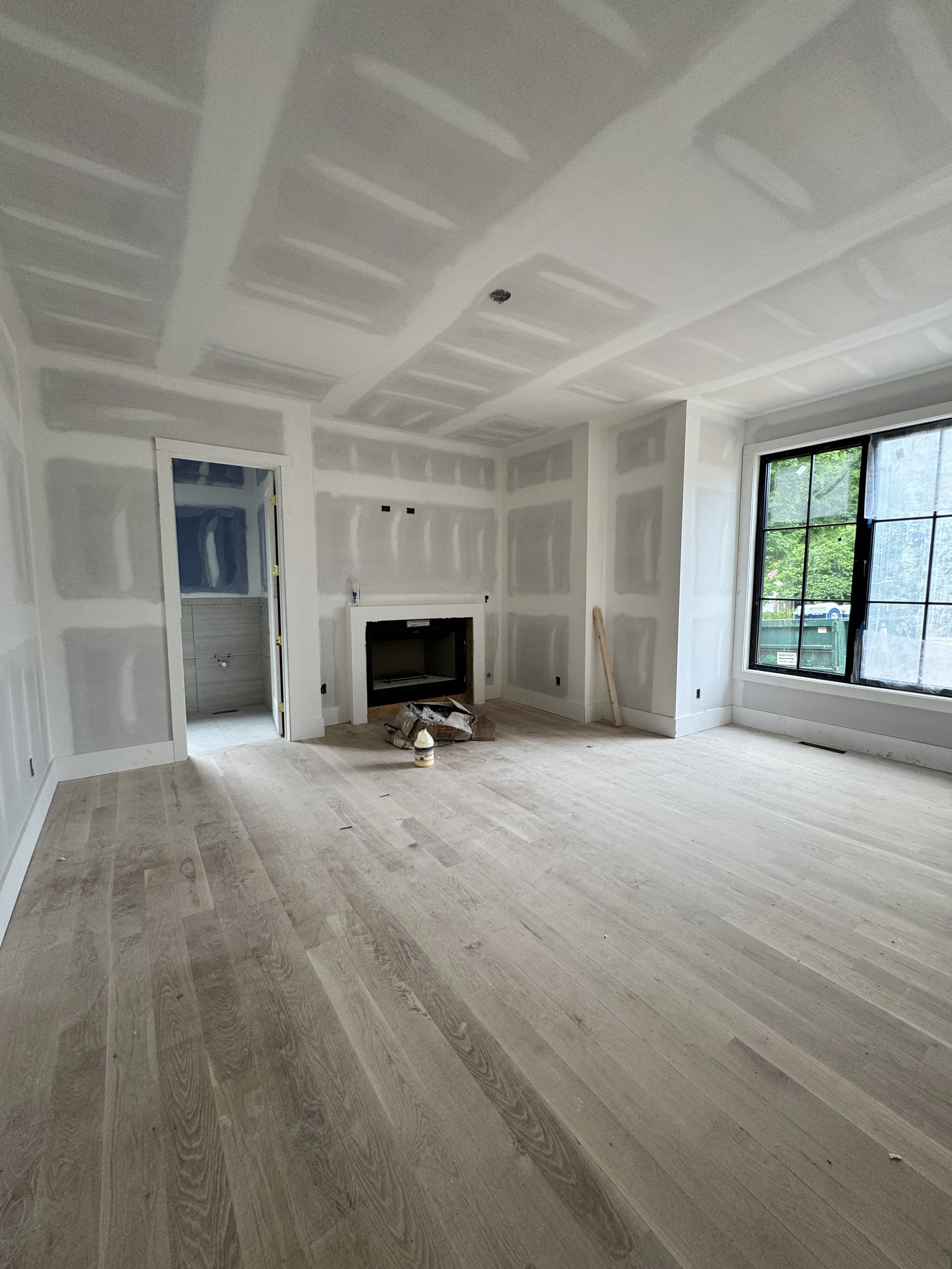 Room under construction with drywall panels installed on the ceiling and walls, and wood flooring in place. A window is visible on the right, and construction materials and tools are on the floor near the fireplace.