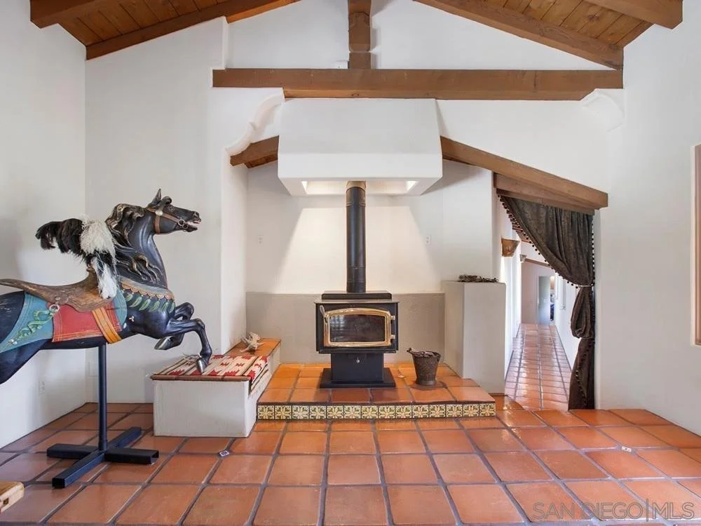 A living room with a brick fireplace, a black wood stove, and a wooden ceiling. There is a decorative horse sculpture on the left, and an open doorway with a dark curtain on the right.