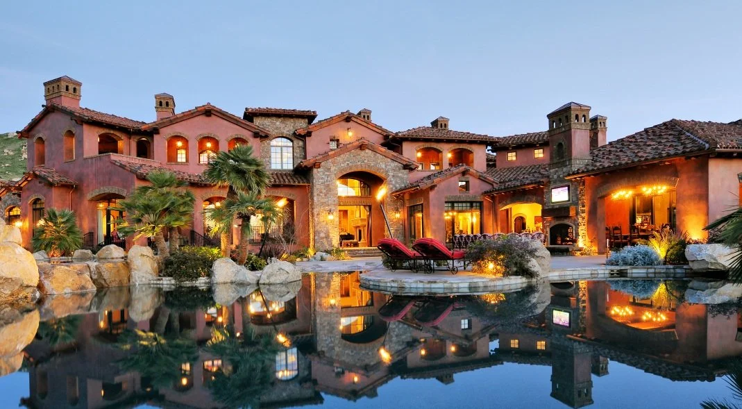 A luxurious house with multiple arches and a tiled roof, surrounded by a landscaped yard with palm trees and rocks, overlooking a reflecting pool at sunset.