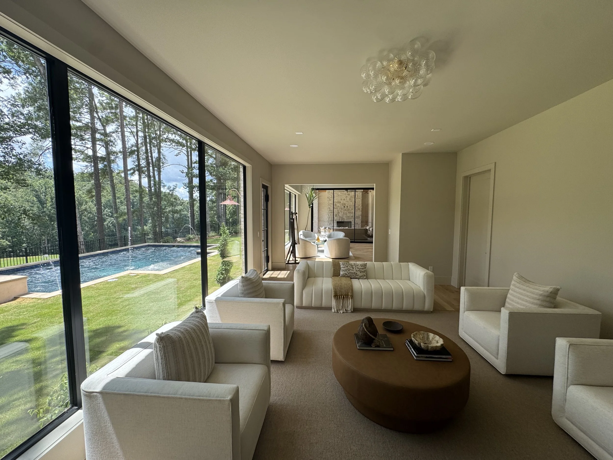Living room with white sofas and a round wooden coffee table, large glass windows overlooking a backyard with a swimming pool and trees, and a sunny outdoor scene.