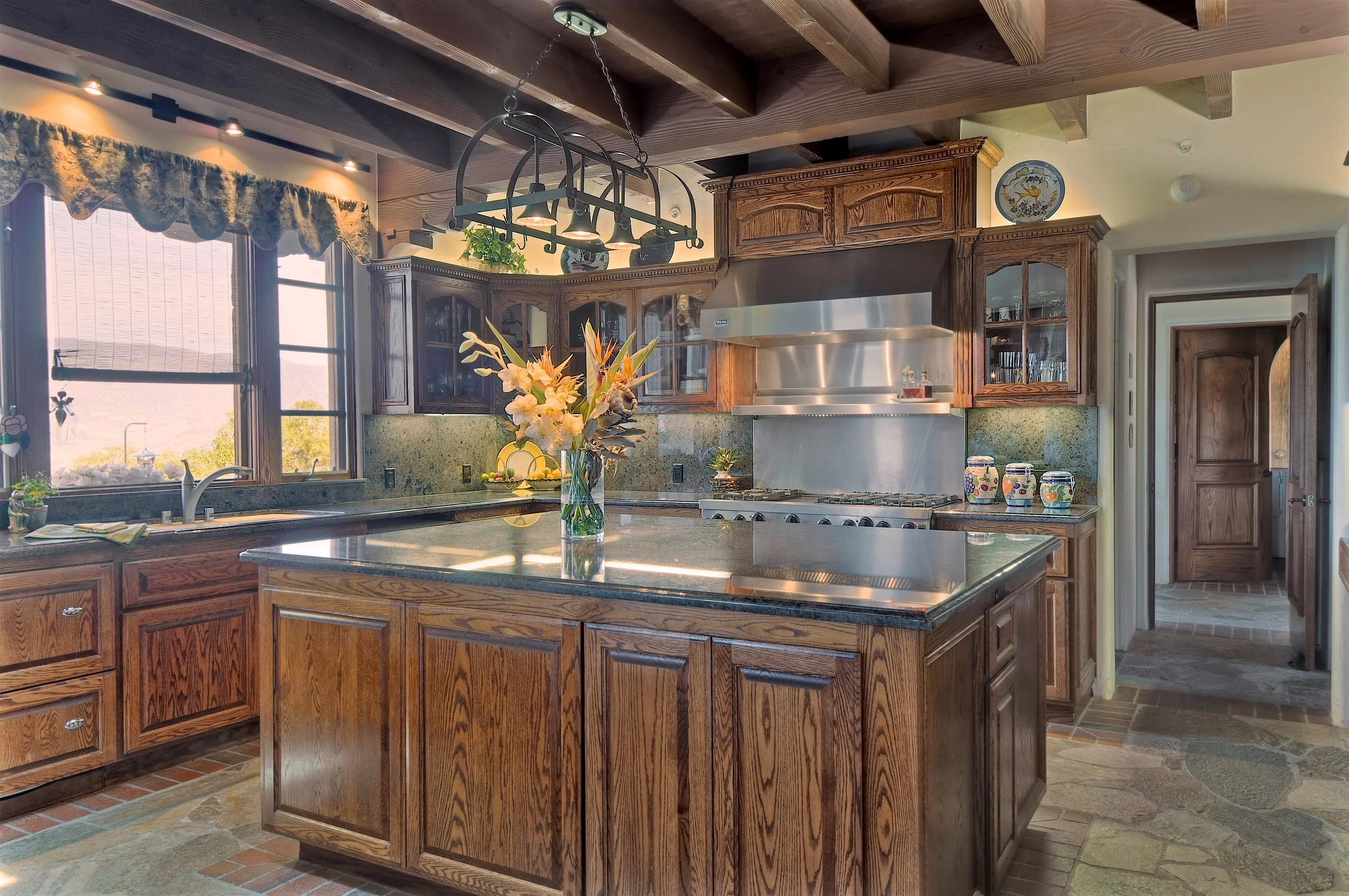 Kitchen with wooden cabinets, a stone countertop, a central island with flowers, large windows with curtains, and a stainless steel range hood, with a chandelier hanging from the ceiling.
