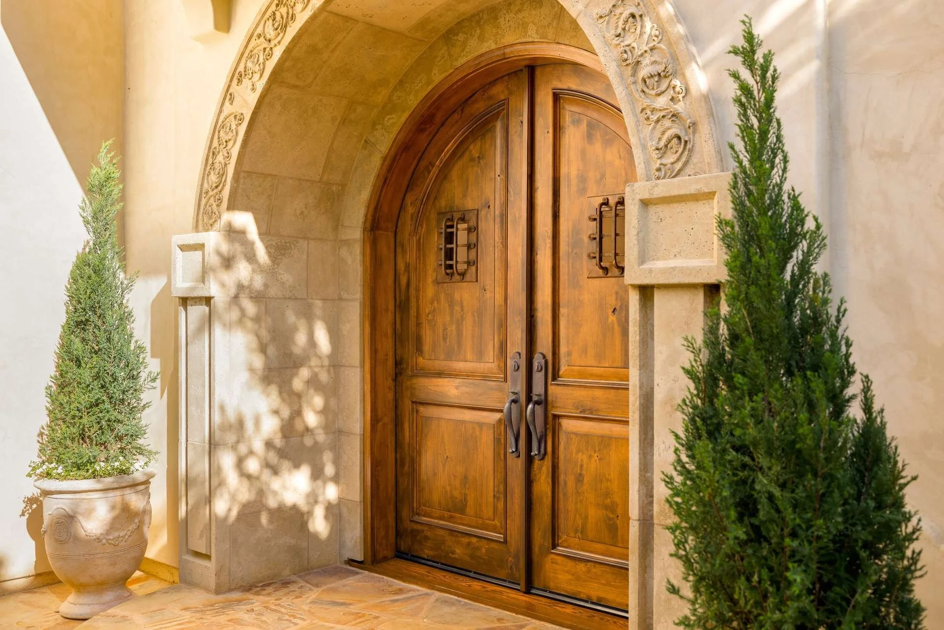 Wooden double front door with vertical panels, black handles, and decorative window grilles, set within a stone archway with ornate carvings, flanked by potted plants, under sunlight.