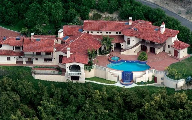 A large luxurious mansion with red tile roofs, surrounded by lush greenery. The property features a curved swimming pool with a waterfall, hot tub, and extensive patio area.