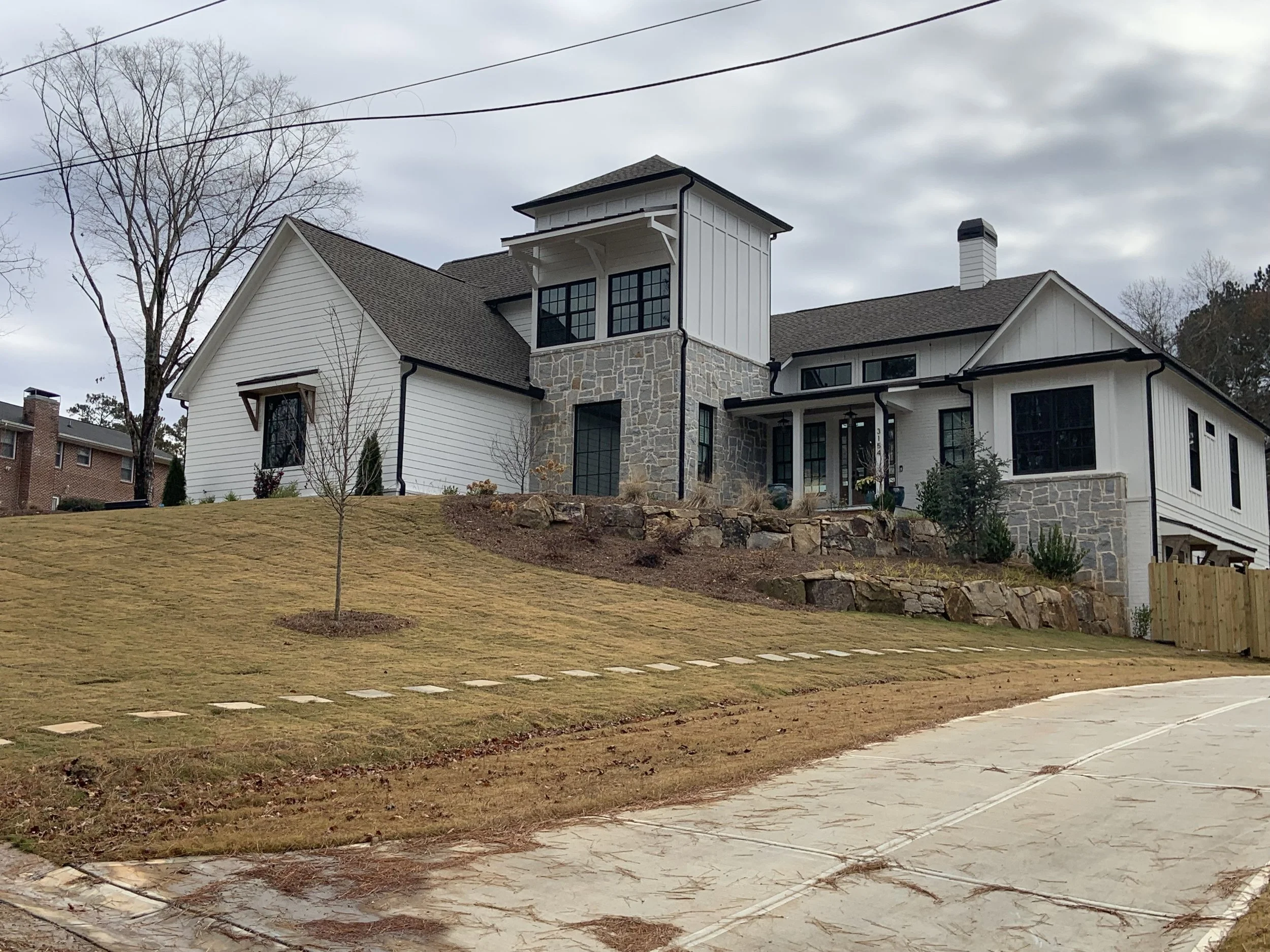 A modern house with white siding, stone accents, and black window frames situated on a lawn with sparse trees and a cloudy sky overhead.