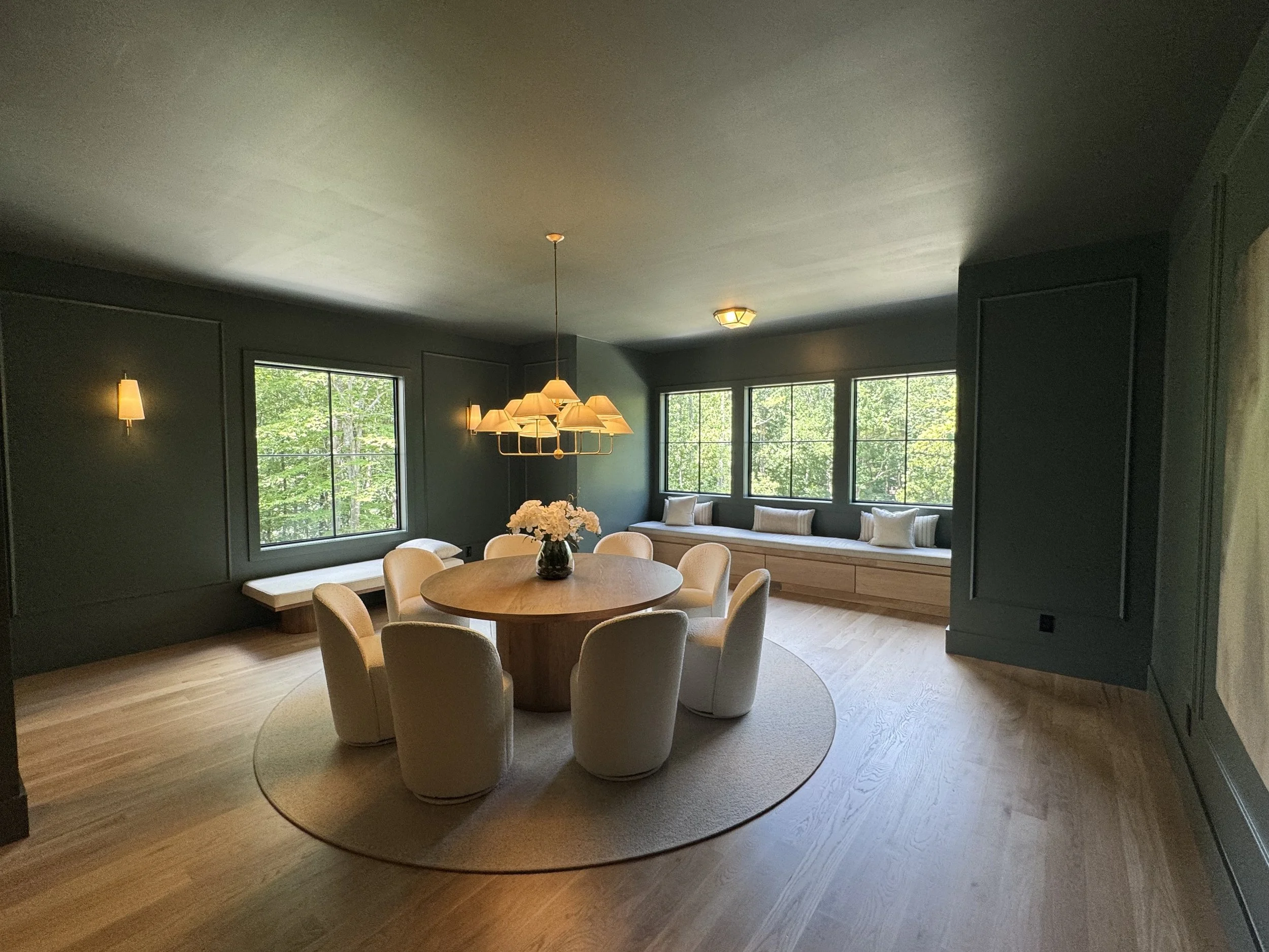 A dining room with a round table, eight beige chairs, a door leading outside, large windows showing trees, and a chandelier hanging above the table.