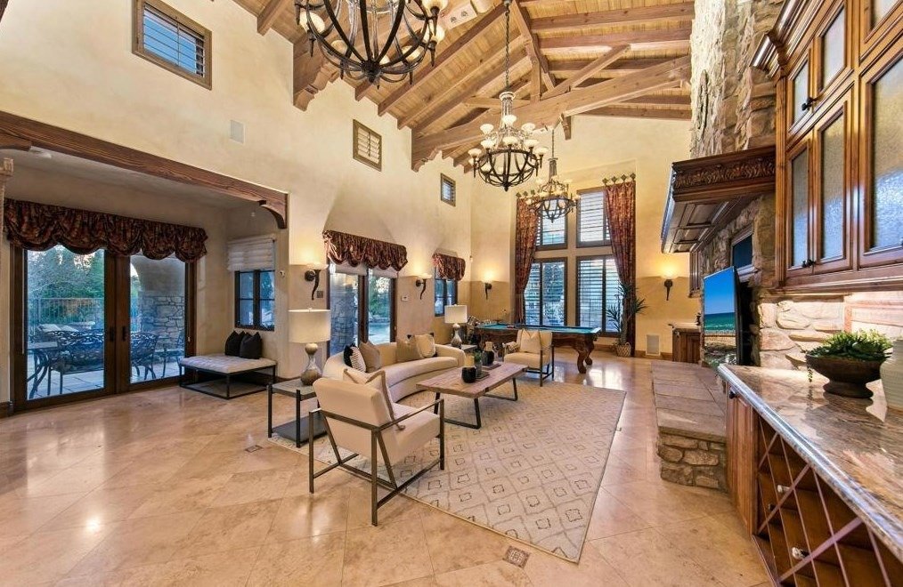 Spacious living room with high vaulted wooden ceiling, large windows with curtains, stone fireplace, chandelier, and comfortable furniture including sofas and chairs.