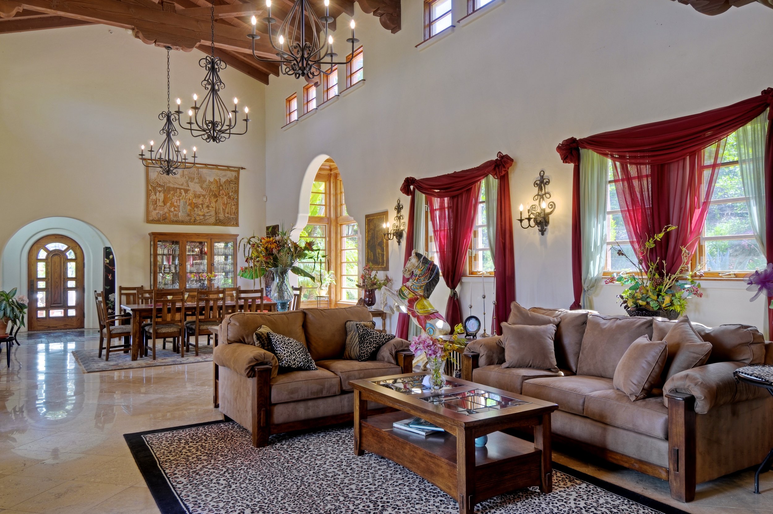 Living room with beige couches, glass-topped coffee table, large windows with red and green curtains, and a high ceiling with wooden beams and chandeliers.