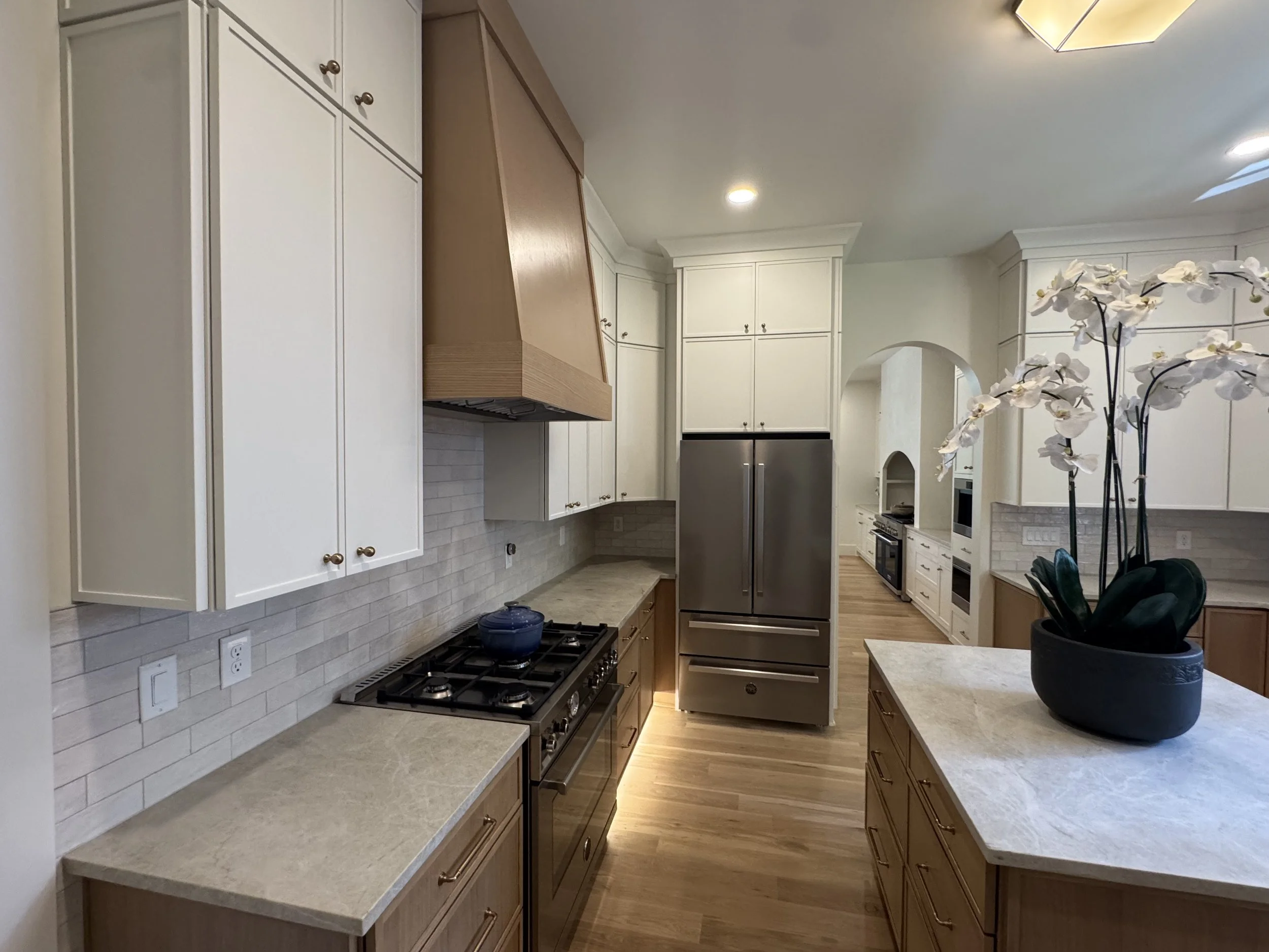 A modern kitchen with white cabinets, a stainless steel refrigerator, a beige countertop, a wood range hood over a stove, a potted orchid on the counter, and recessed ceiling lights.