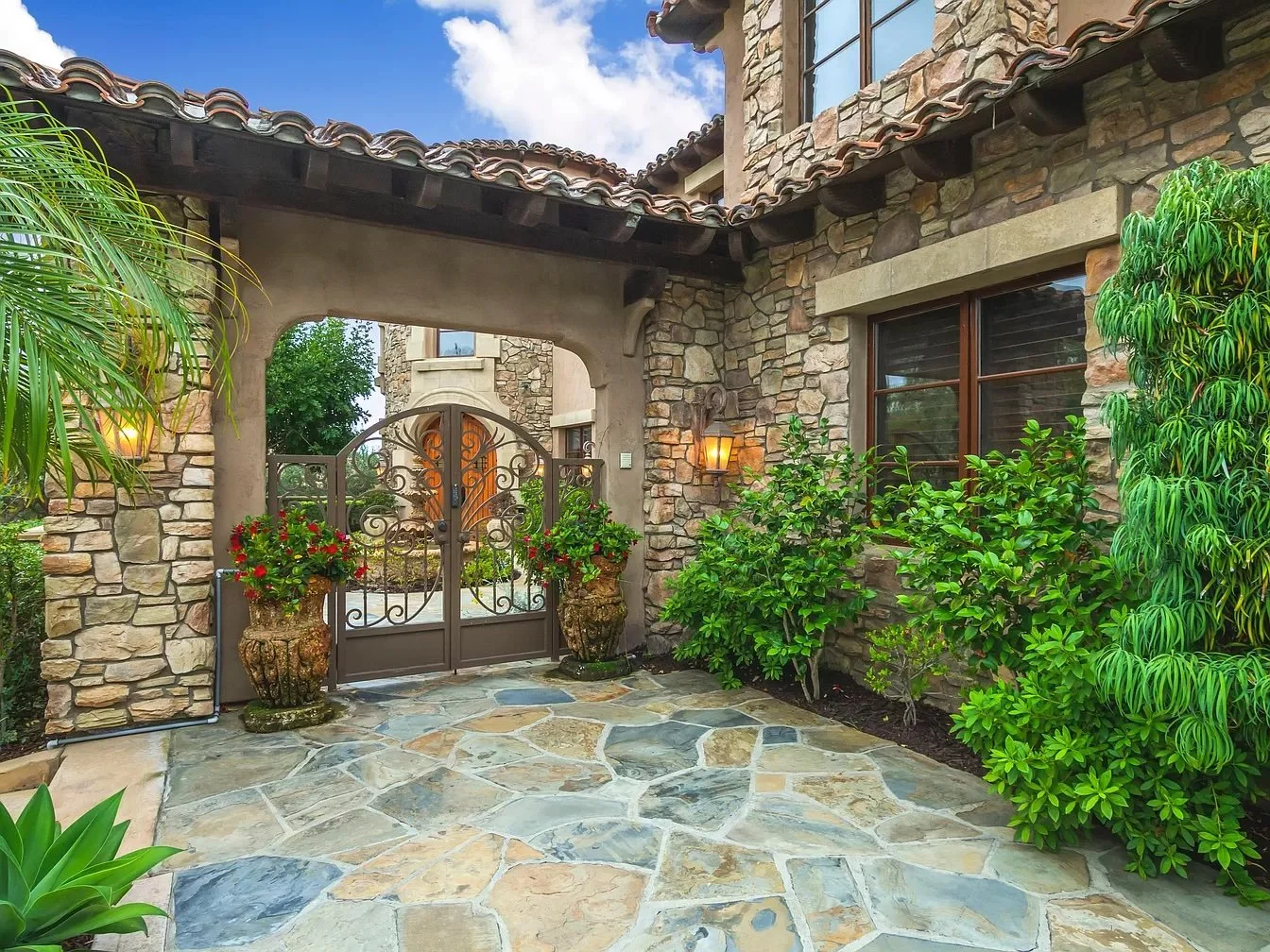 Stone house entrance with wrought iron gate, potted plants, lush greenery, and stone pathway leading to the door, under a tiled roof with outdoor lighting.