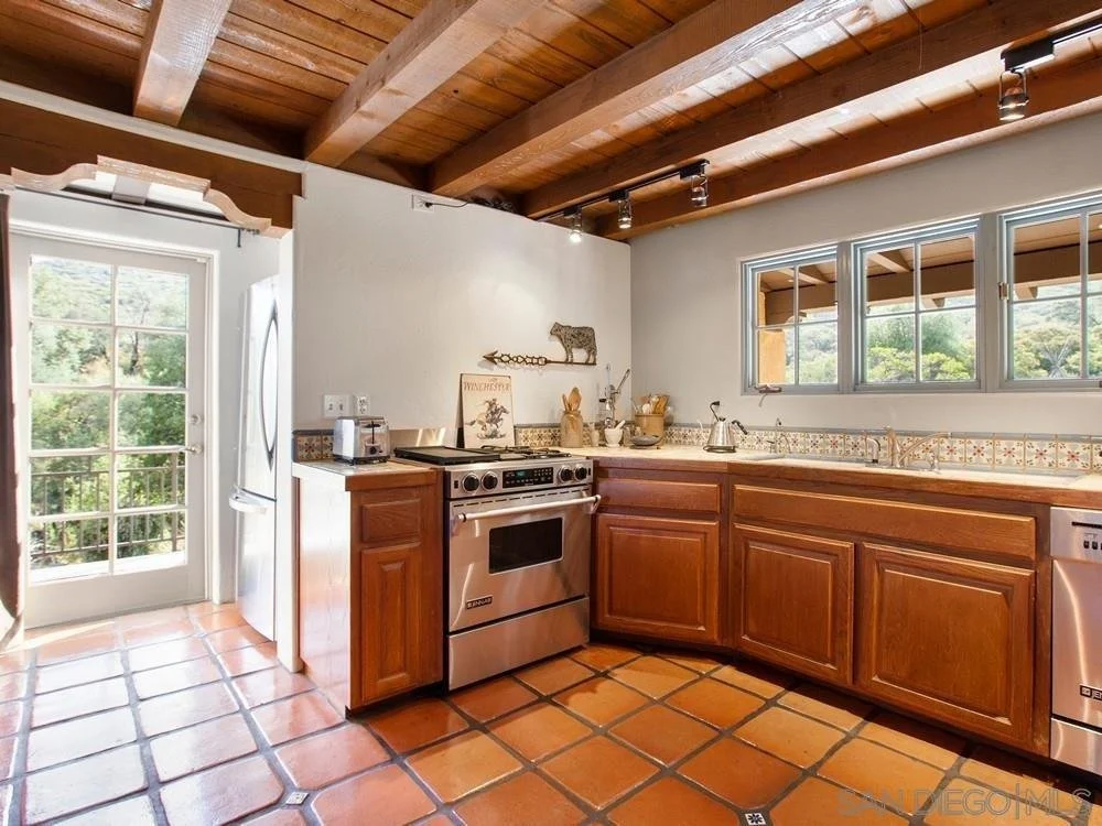 Kitchen with wooden cabinets, terracotta tile floor, stainless steel stove, microwave, and dishwasher, with windows showing greenery outside and a glass door leading outside.